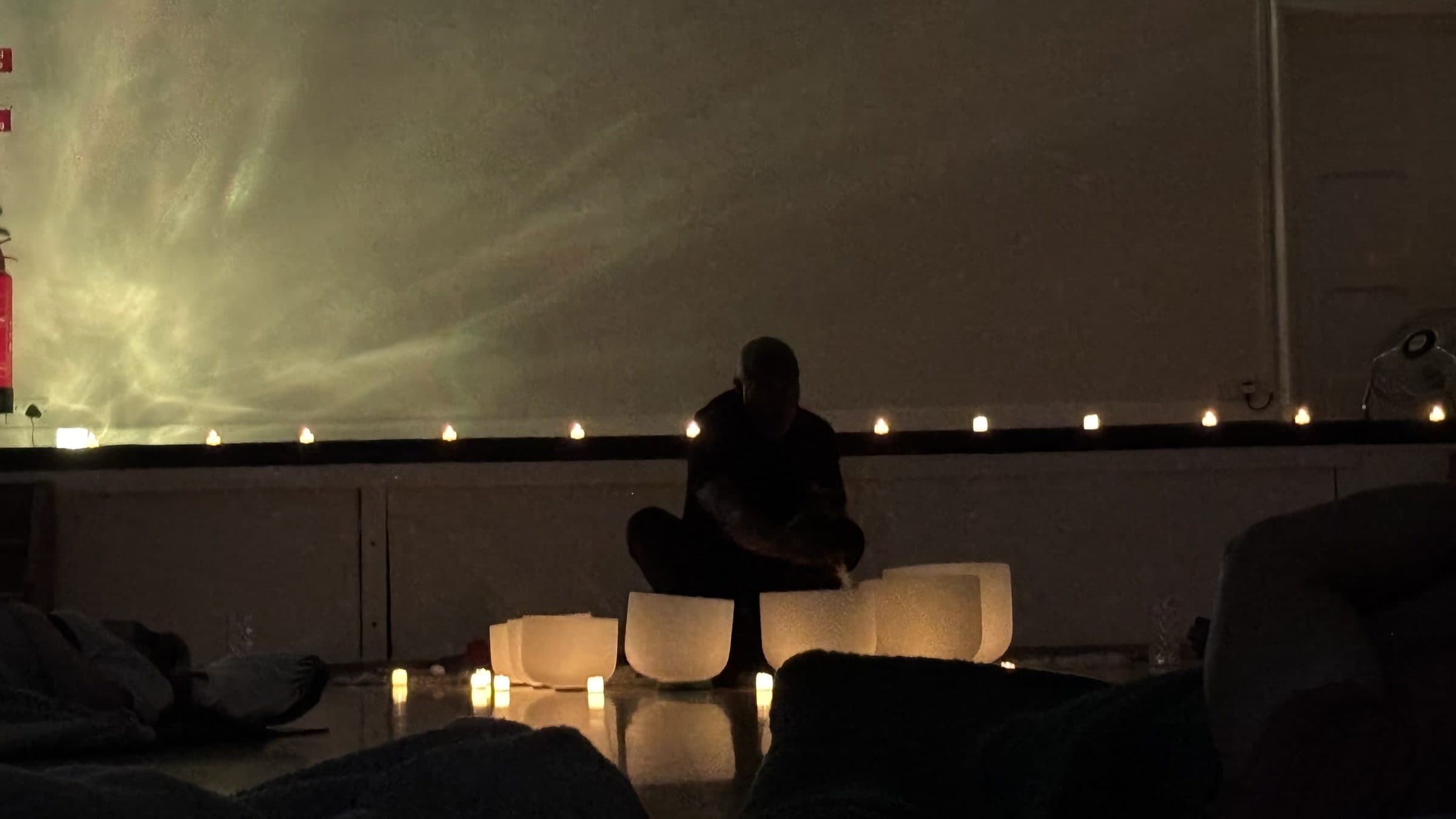 Clinton playing the crystal singing bowls by candlelight at the front of the hall, with people resting on mats in the foreground