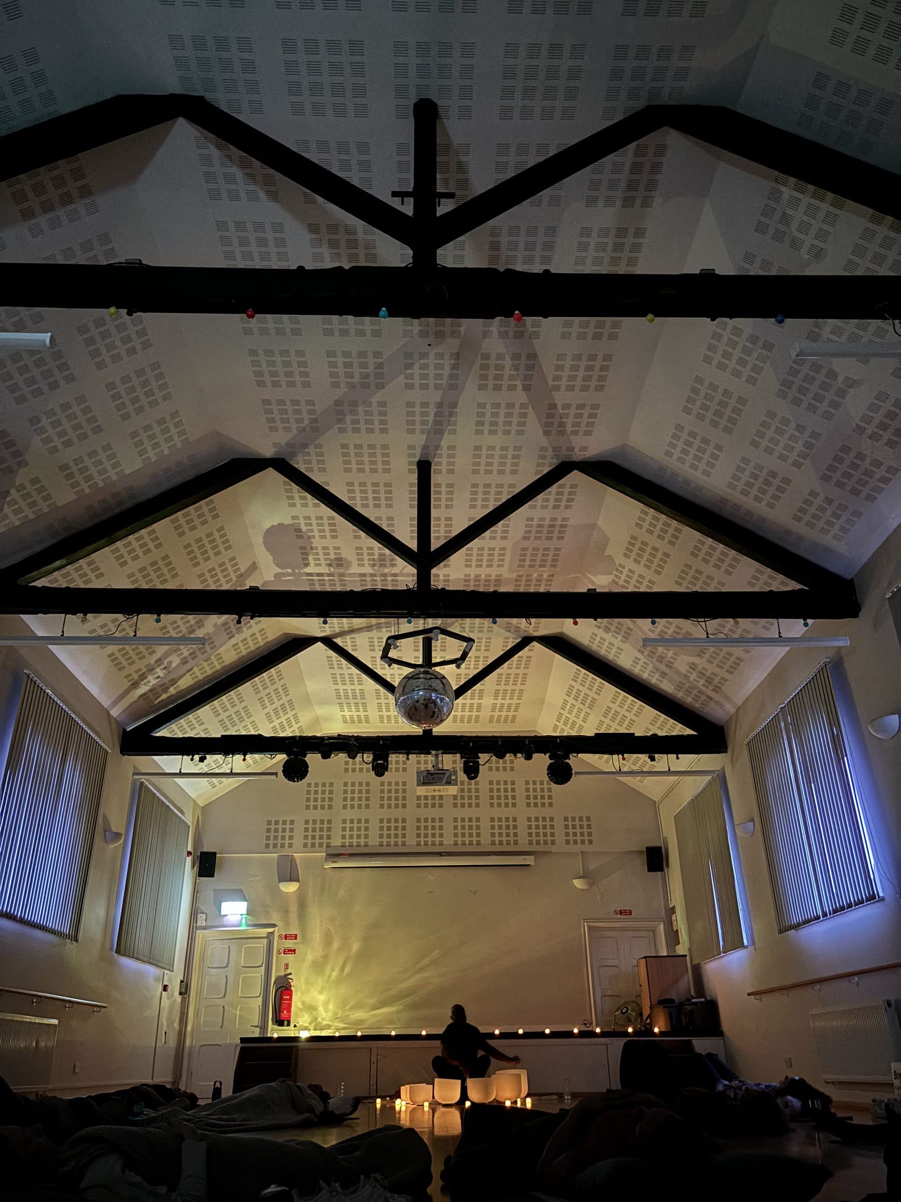 The beamed ceiling and mirror ball of the village hall above the session, with people lying on mats below