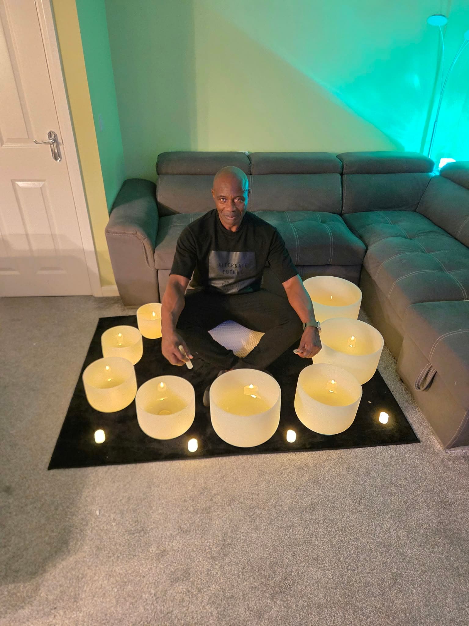 Eight crystal singing bowls arranged in a circle on a black mat, each lit by a candle, with Clinton seated in the middle